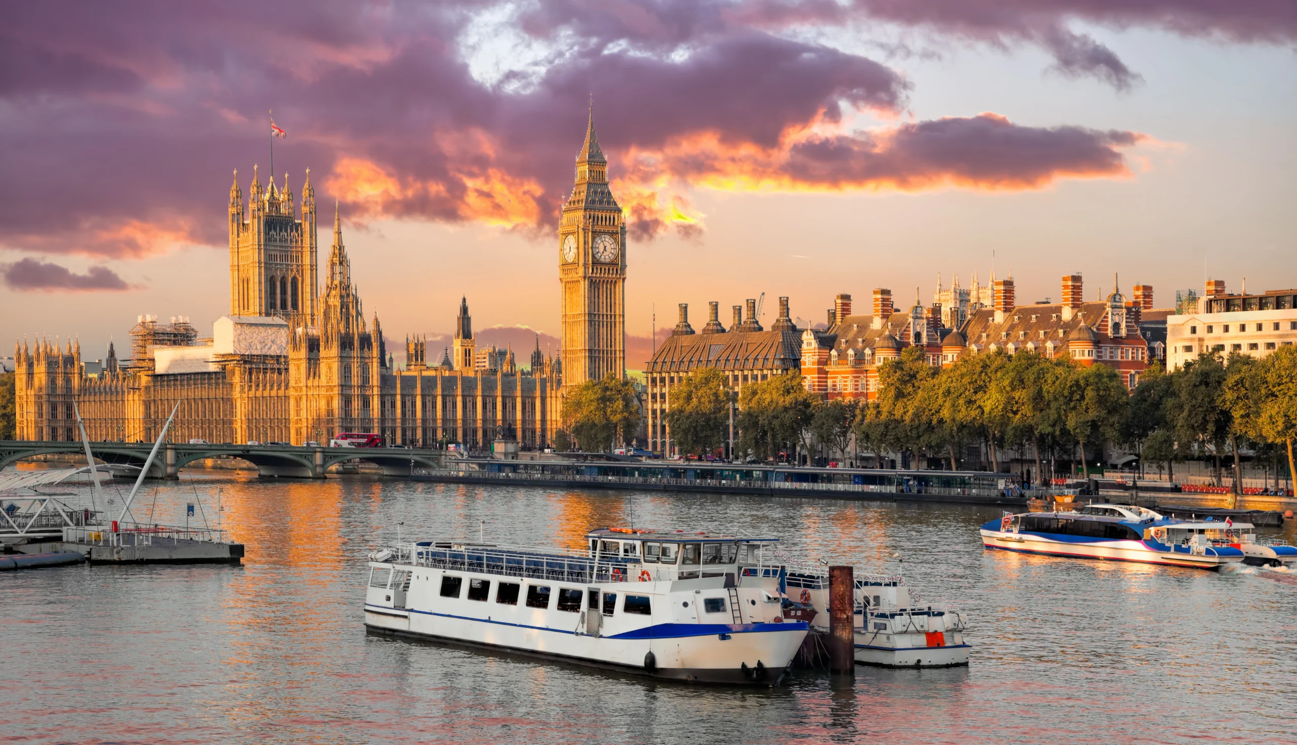 Sunset over the London Thames waterfront featuring Big Ben and the Houses of Parliament with moored boats.